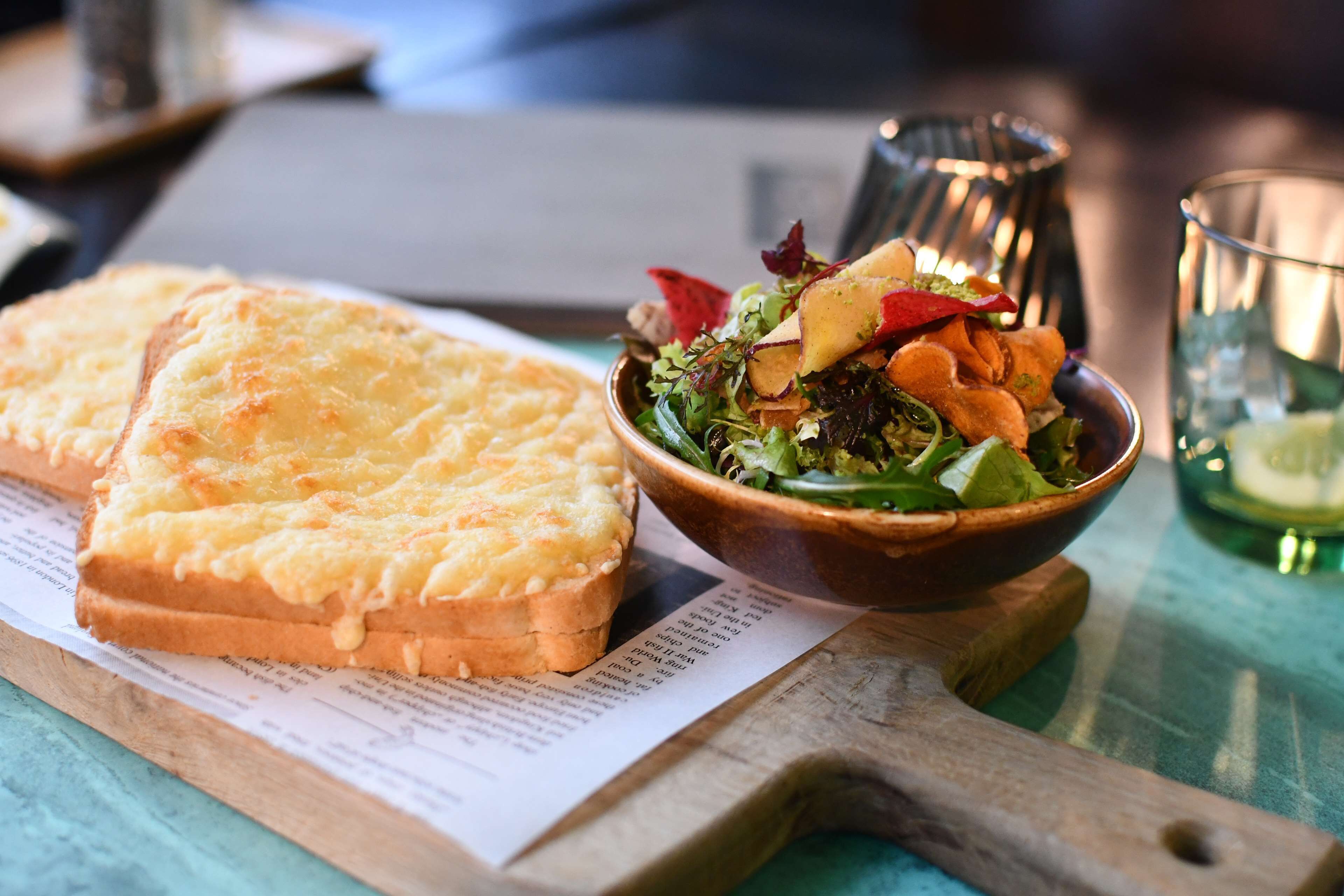 A wooden cutting board with a piece of toasted bread and a bowl of salad on a table with a menu.