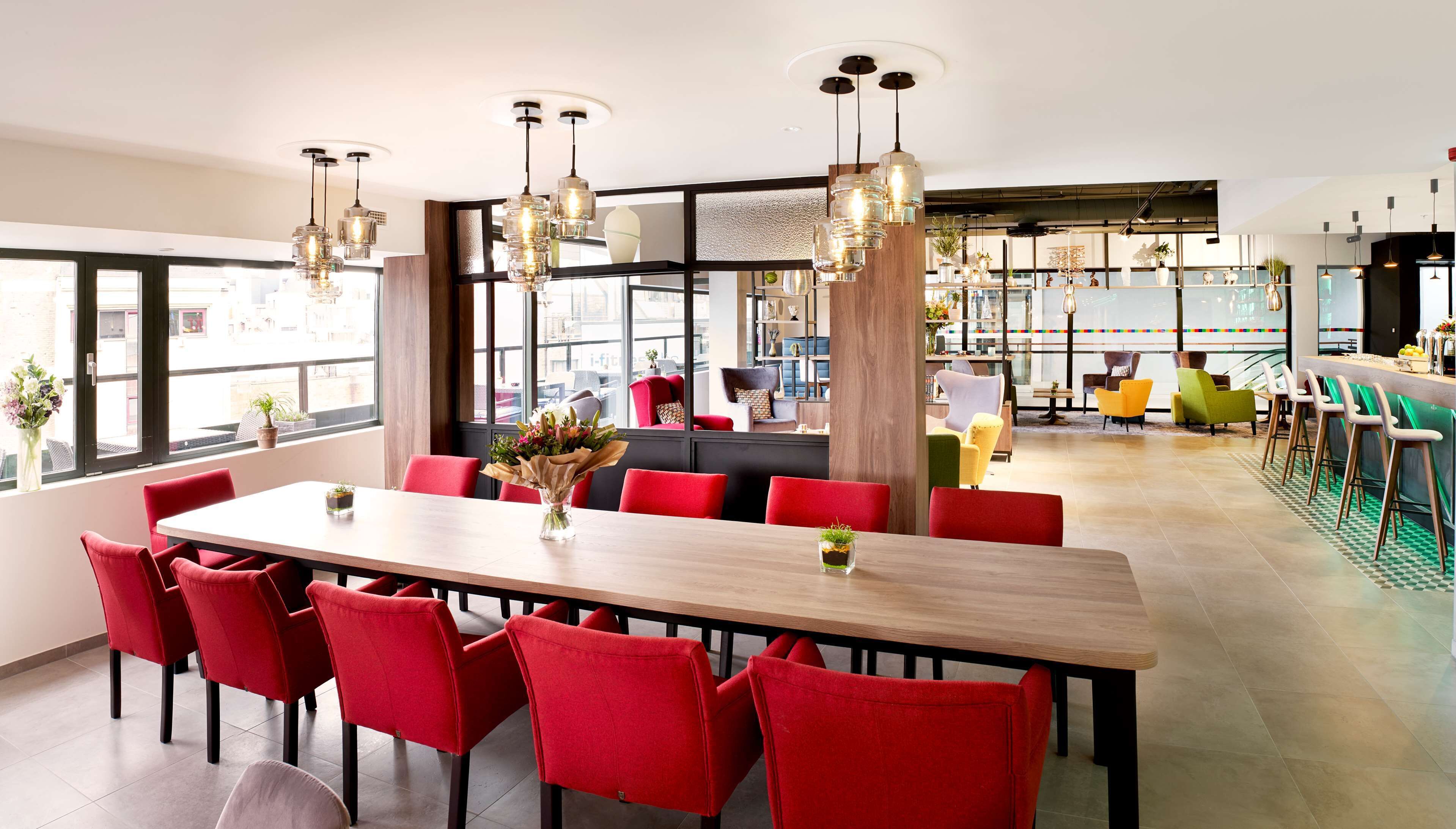 A modern dining area with a long wooden table, red upholstered chairs, and hanging pendant lights.