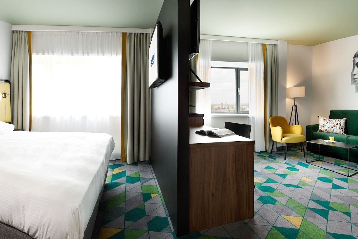 Habitación de hotel con cama, escritorio, sillón, sofá y un amplio ventanal con cortinas.