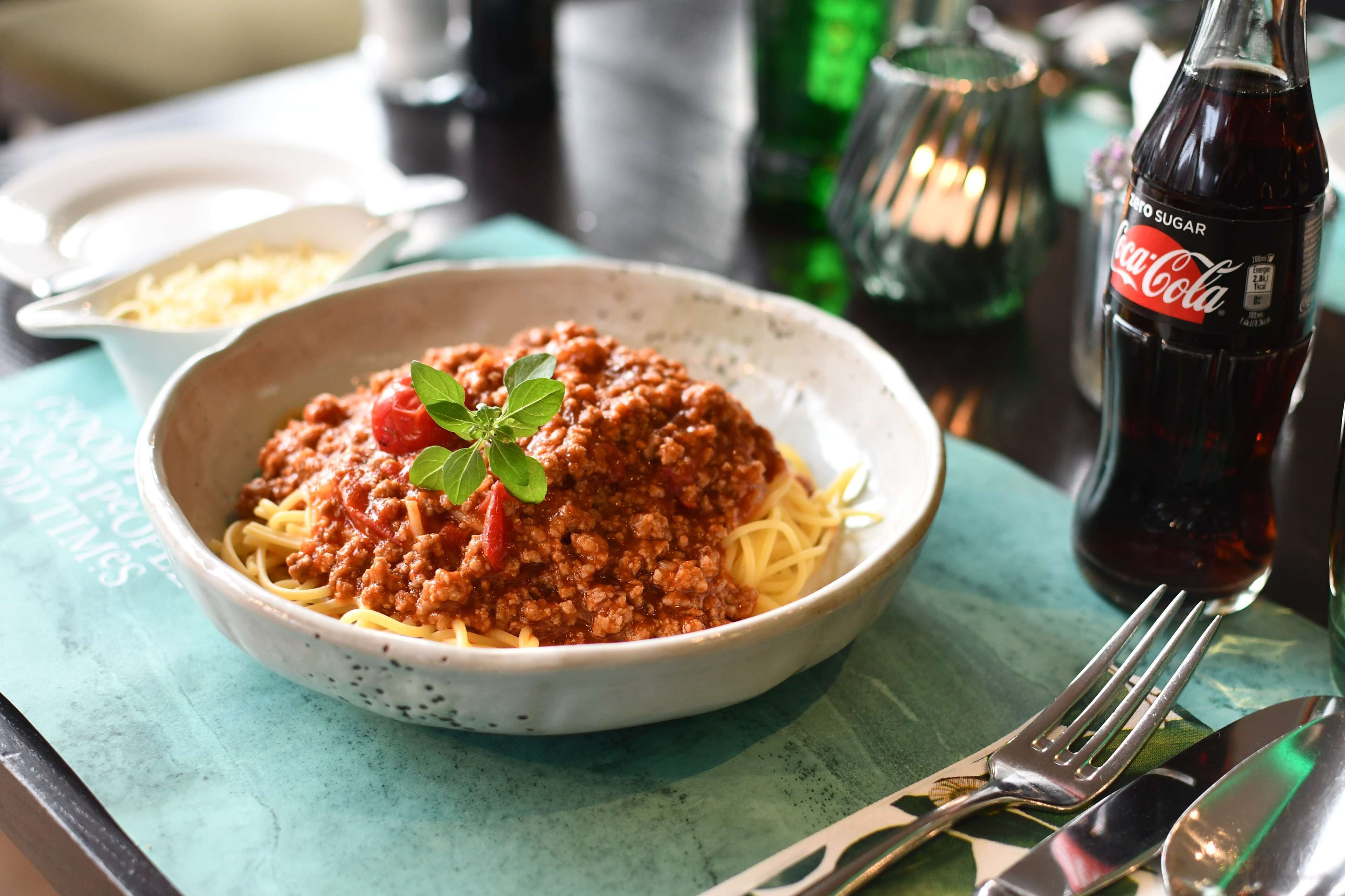 A bowl of spaghetti with meat sauce and a sprig of basil, a Coca-Cola bottle, and cutlery on a table.