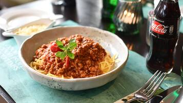 A bowl of spaghetti with meat sauce and a sprig of basil, a Coca-Cola bottle, and cutlery on a table.