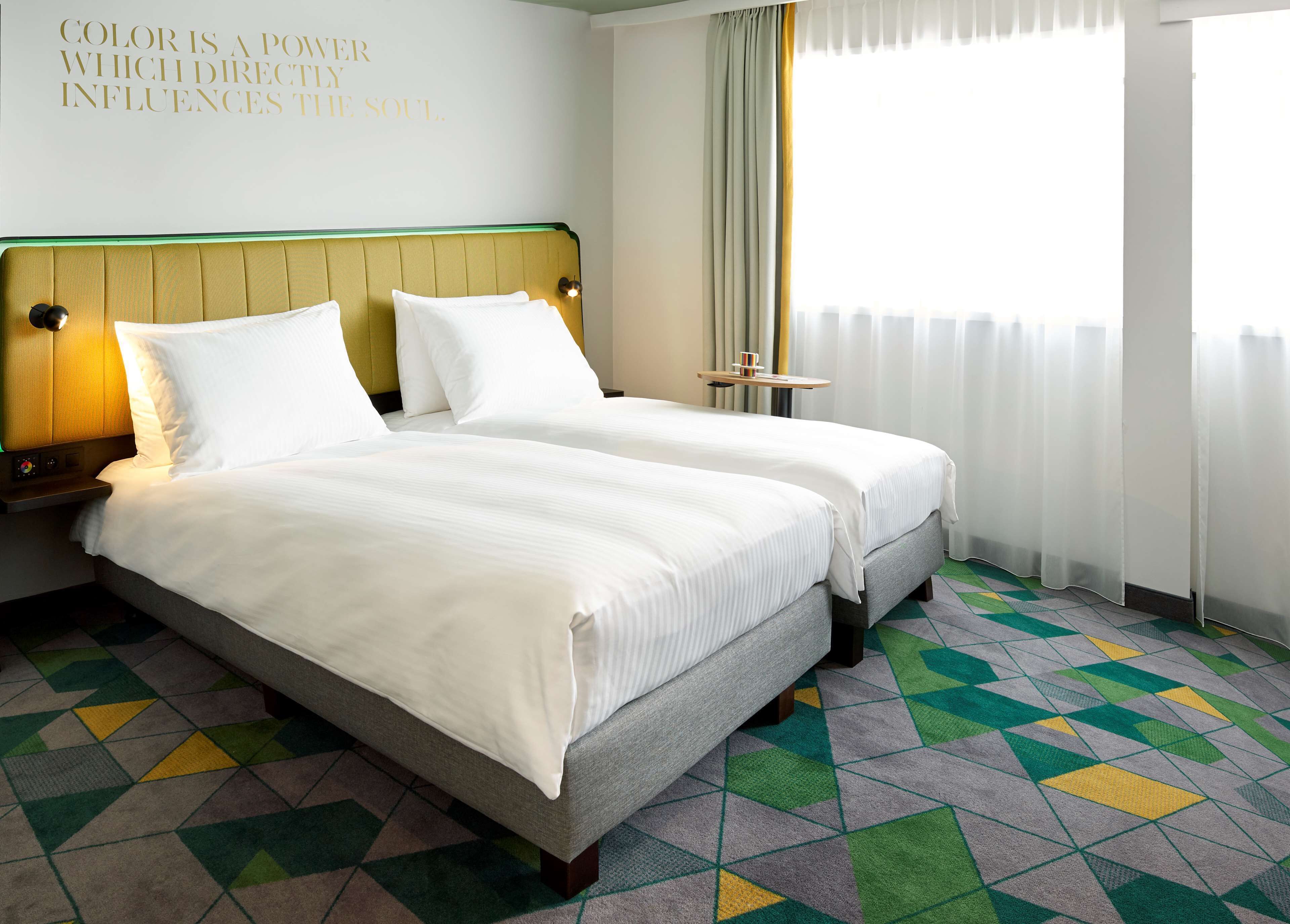 Een hotelkamer met twee bedden, een hoofdeinde, nachtkastjes, een raam met gordijnen en een kamerbreed tapijt met dessin.
