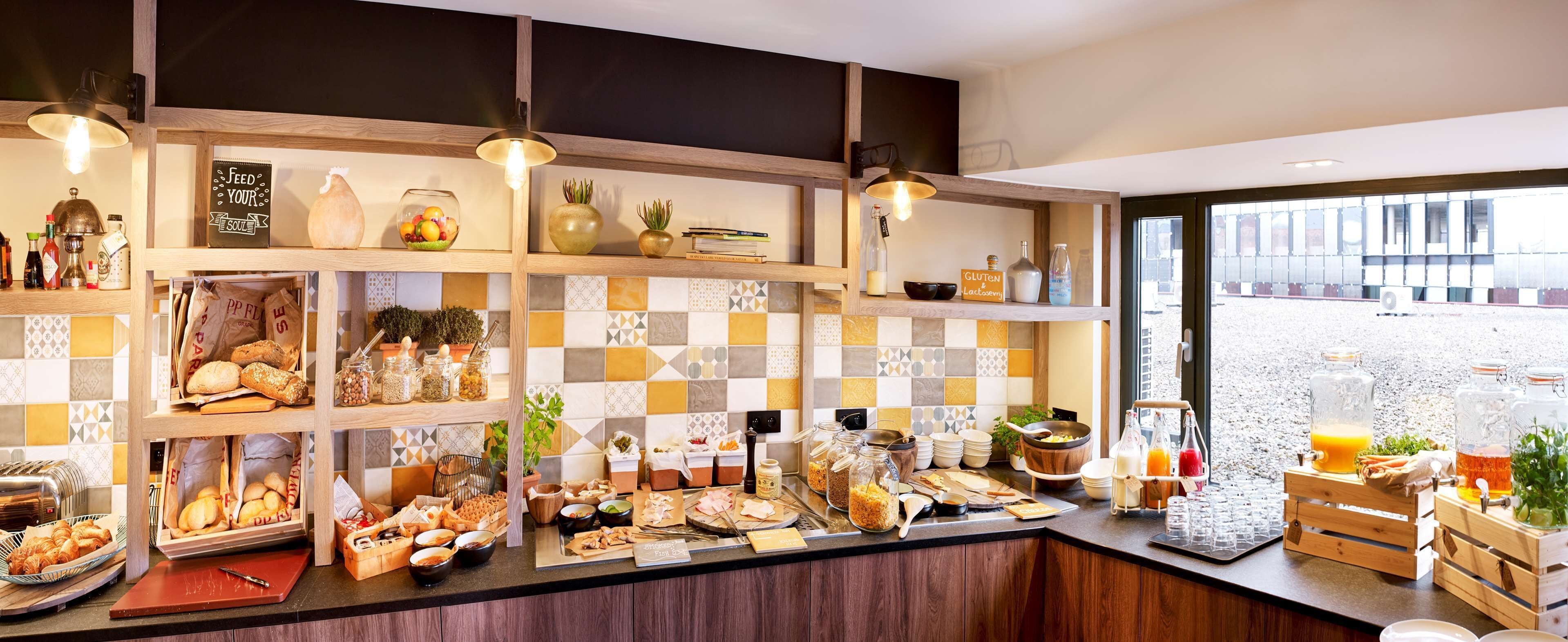 A breakfast buffet with various food items and beverages, wooden shelves, and a checkered tile backsplash.