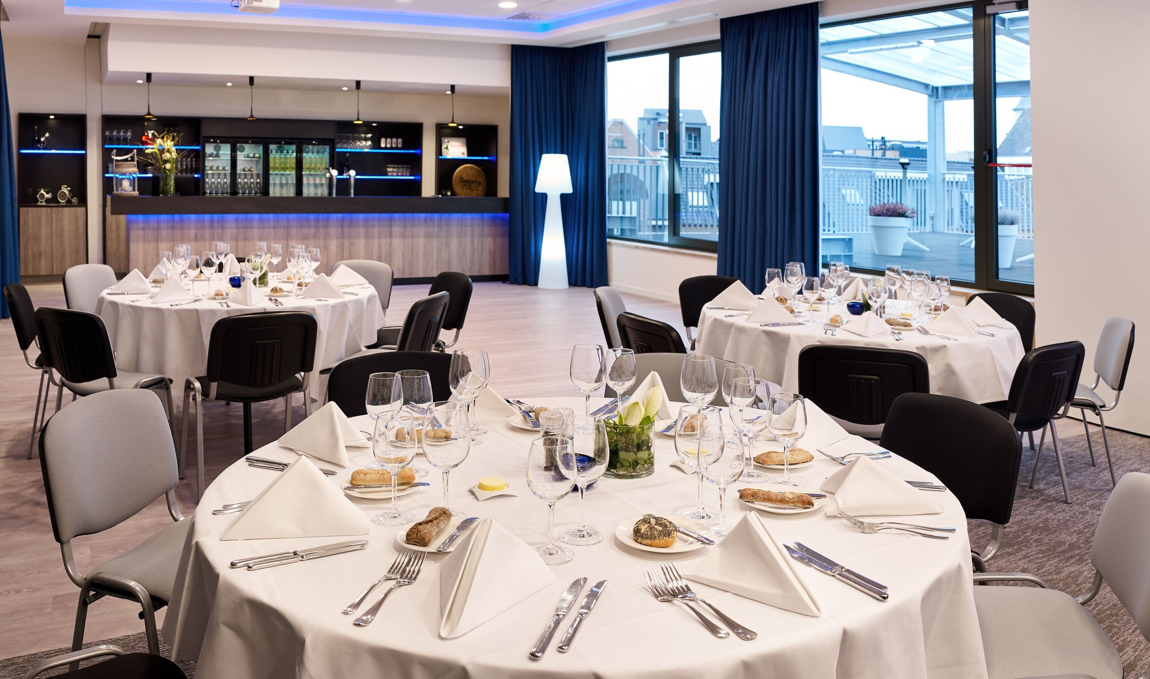 A hotel dining area with round tables set for a meal, a bar with a view, and a large window with curtains.