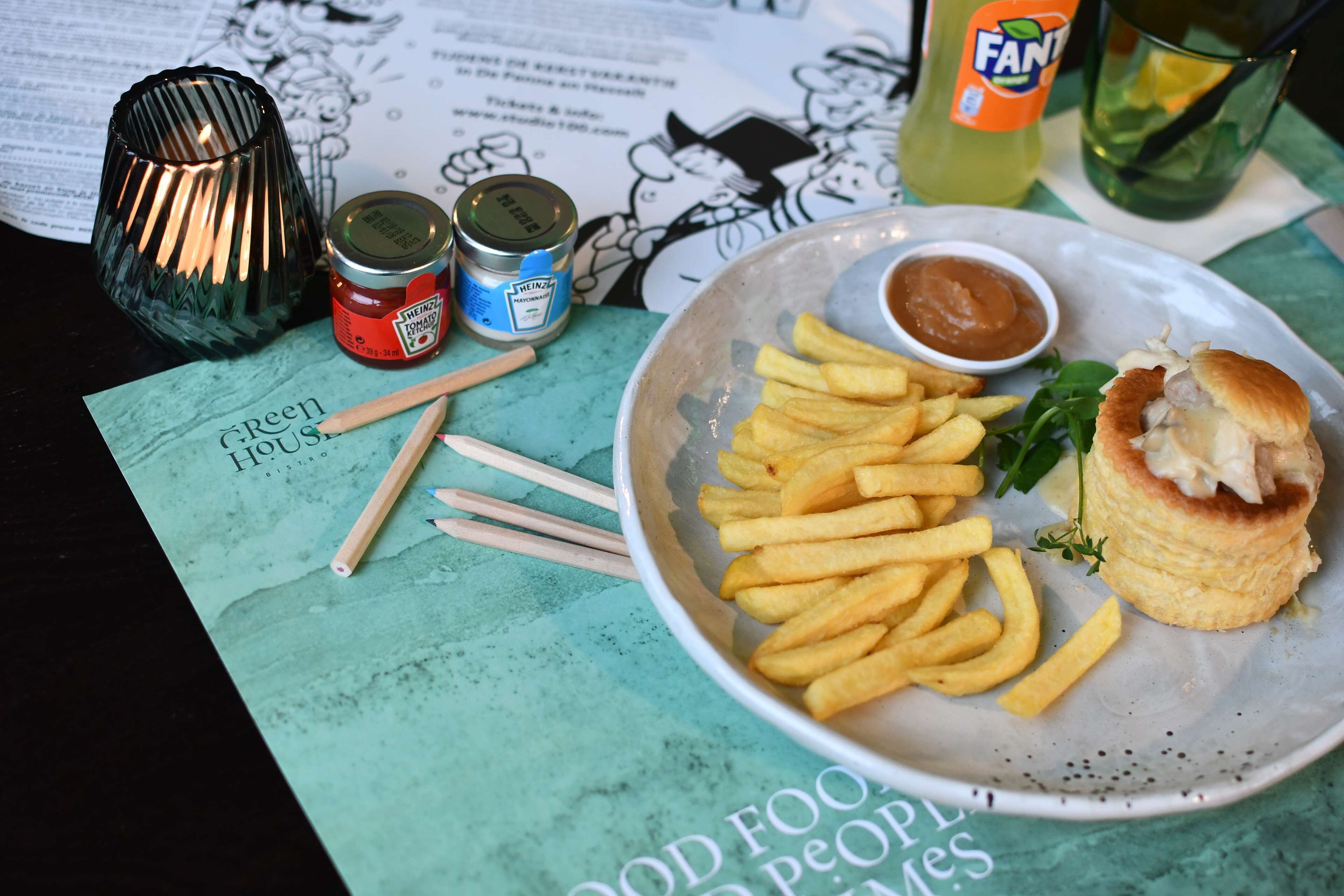 Um prato com batatas fritas e um hambúrguer, uma lata de ketchup Heinz, uma garrafa de Fanta e um menu verde com ilustrações de comida.