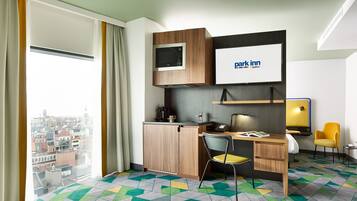 Ett hotellrum med utsikt över staden, ett skrivbord med en stol, en mikrovågsugn, ett litet pentry, en TV och en säng.