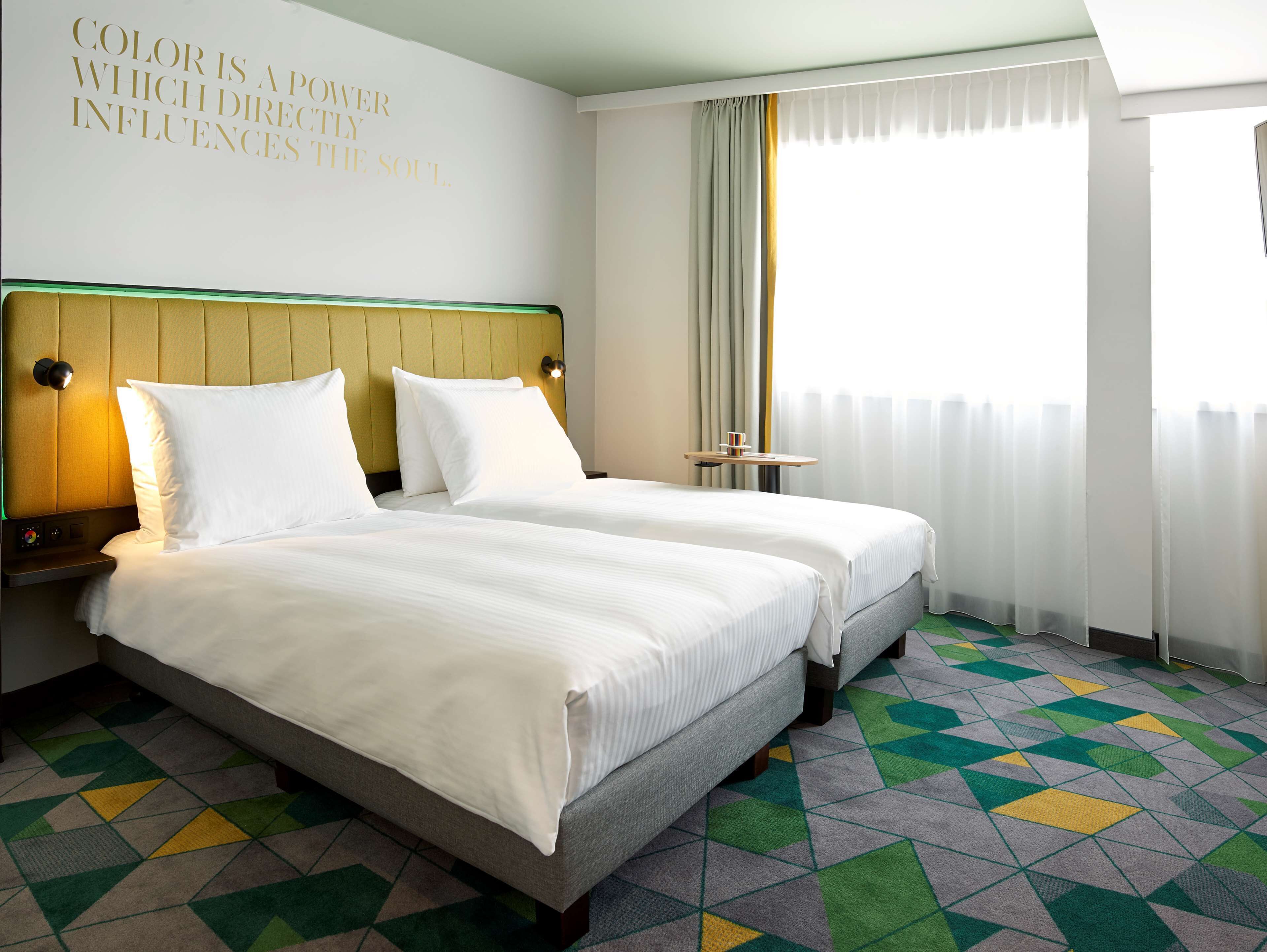 A hotel room with a bed, bedside tables, a wall with a motivational quote, a window with curtains, and a patterned carpet.
