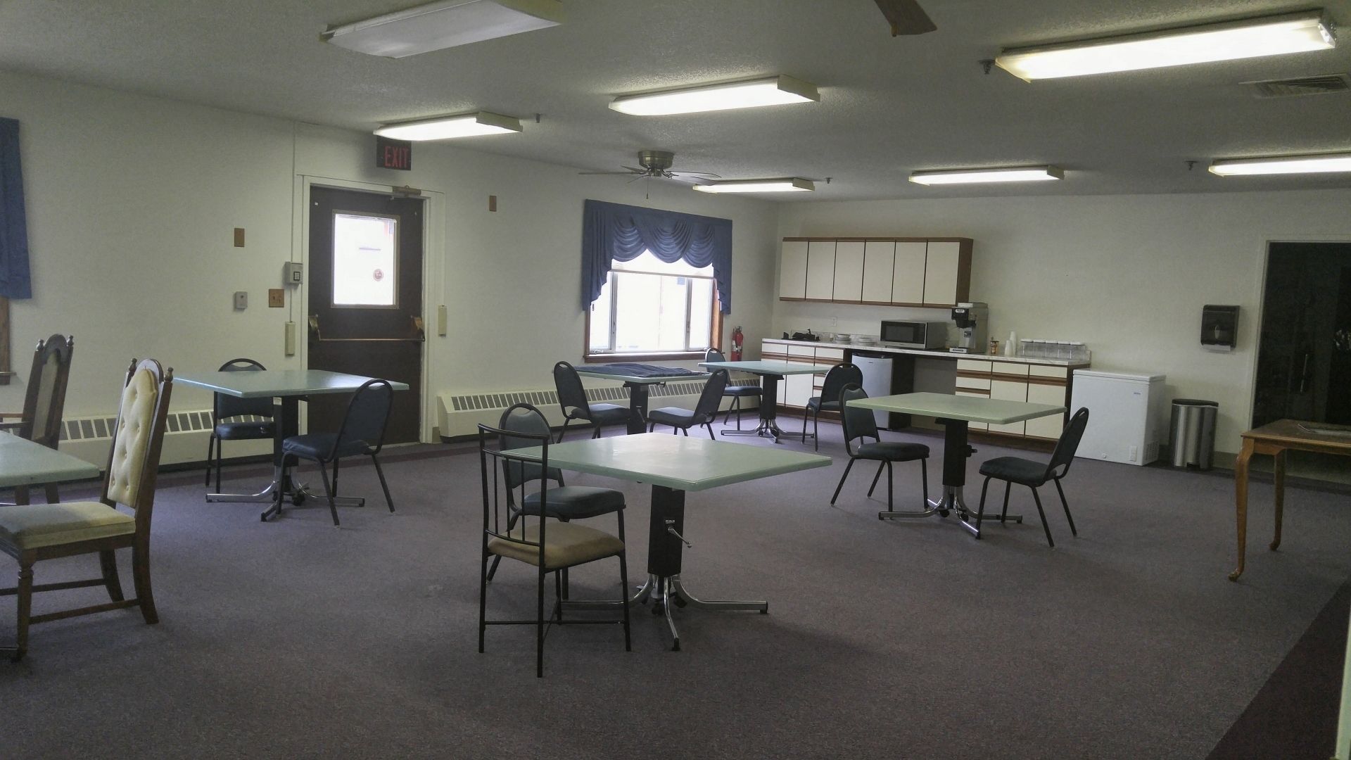 A conference room with multiple tables and chairs, a kitchenette, and an exit door.