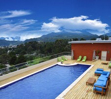 Una piscina en la azotea con terraza de madera, sillones reclinables verdes y vistas a montañas y un pueblo al fondo.