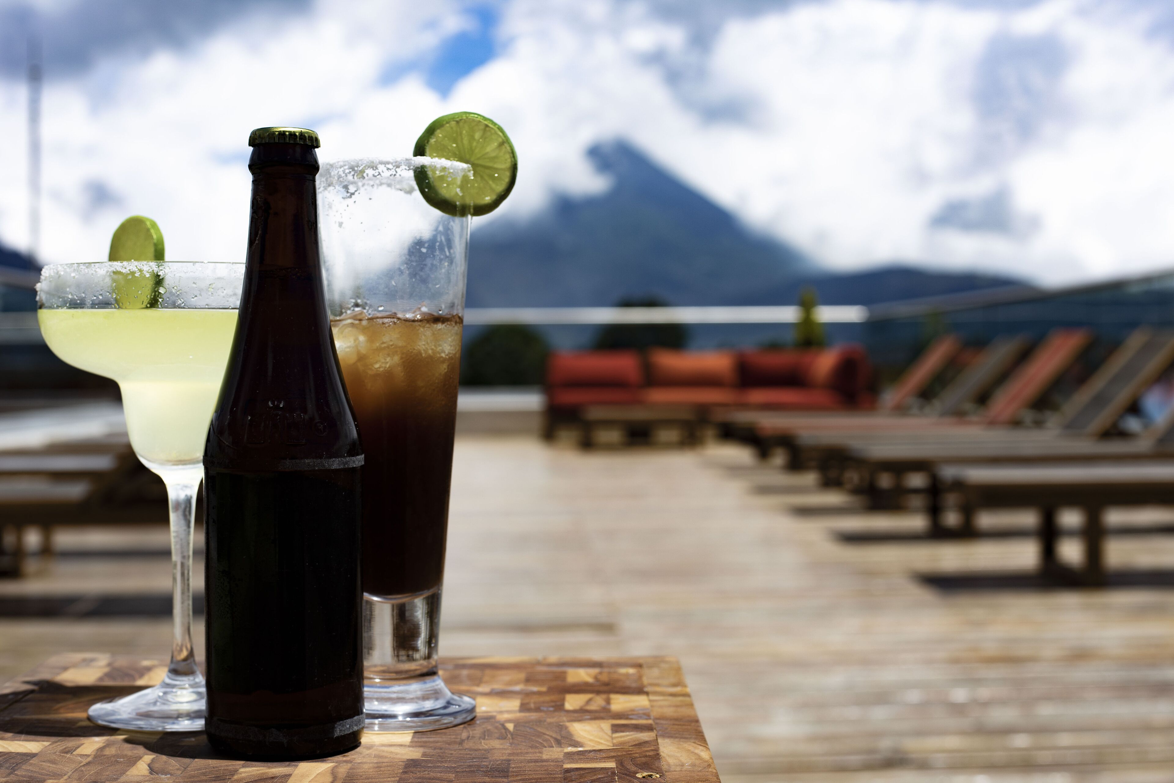 A wooden table with two drinks, one with a lime wedge, and a bottle of beer. The background features a deck with lounge chairs and a mountain view.