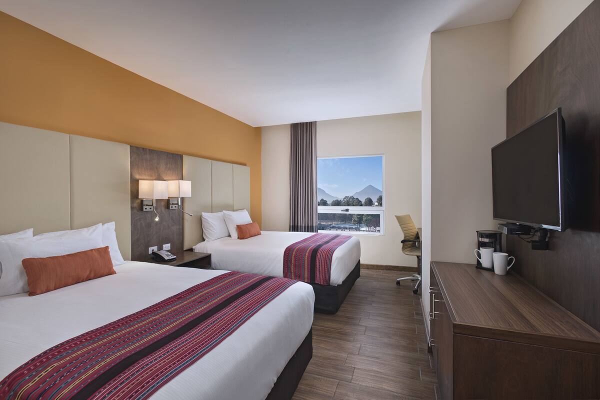 Habitación de hotel con dos camas, un televisor, un escritorio y ventana con vista a la montaña.