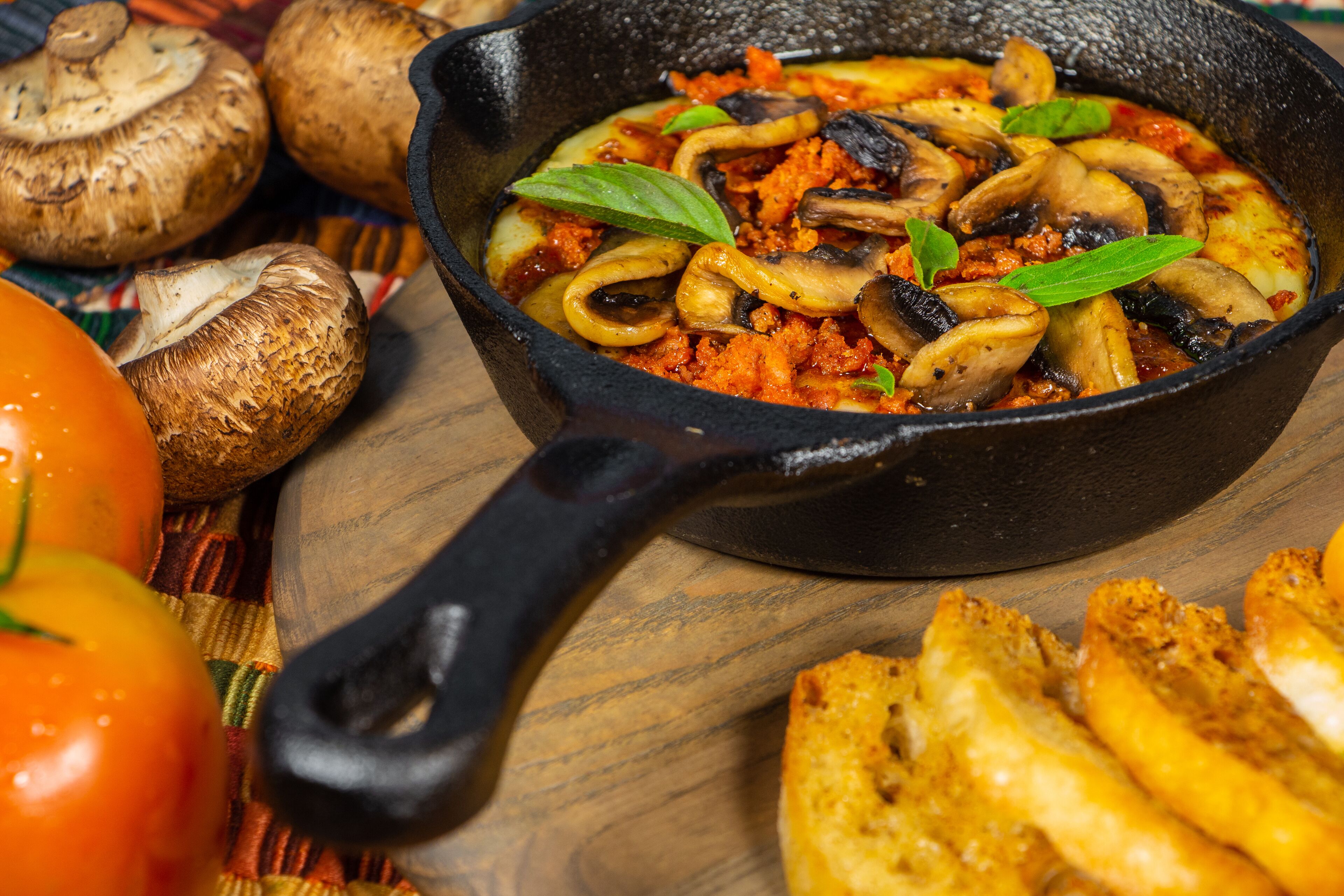 Una sartén con un plato de champiñones y tomates, rodeada de champiñones enteros y un tomate sobre una superficie de madera.