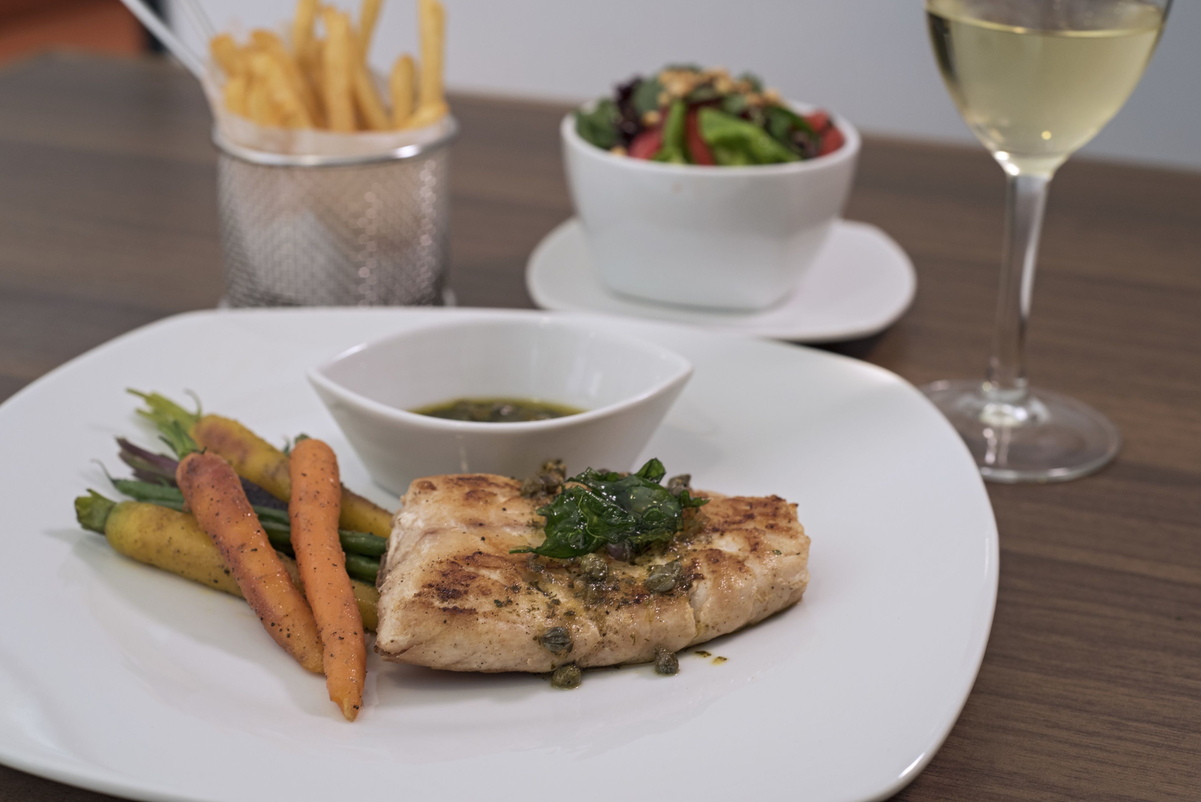 A plate with grilled fish, carrots, and a small bowl of sauce, accompanied by a glass of white wine and a side salad.