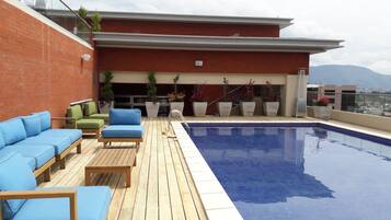 Un área de piscina en la azotea con sillones azules y verdes, una terraza de madera y plantas en macetas.