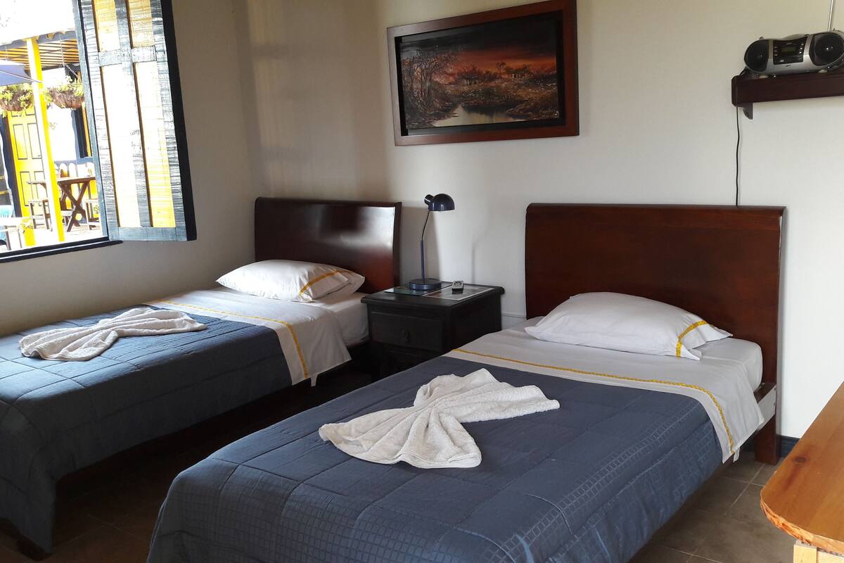 A hotel room with two single beds, a nightstand with a lamp, a painting on the wall, and a window with a view of the outside.