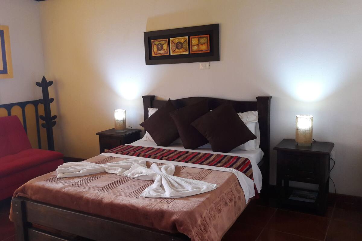 A neatly made bed with a striped bedspread, two bedside lamps, a red sofa, and a wall-mounted picture frame.