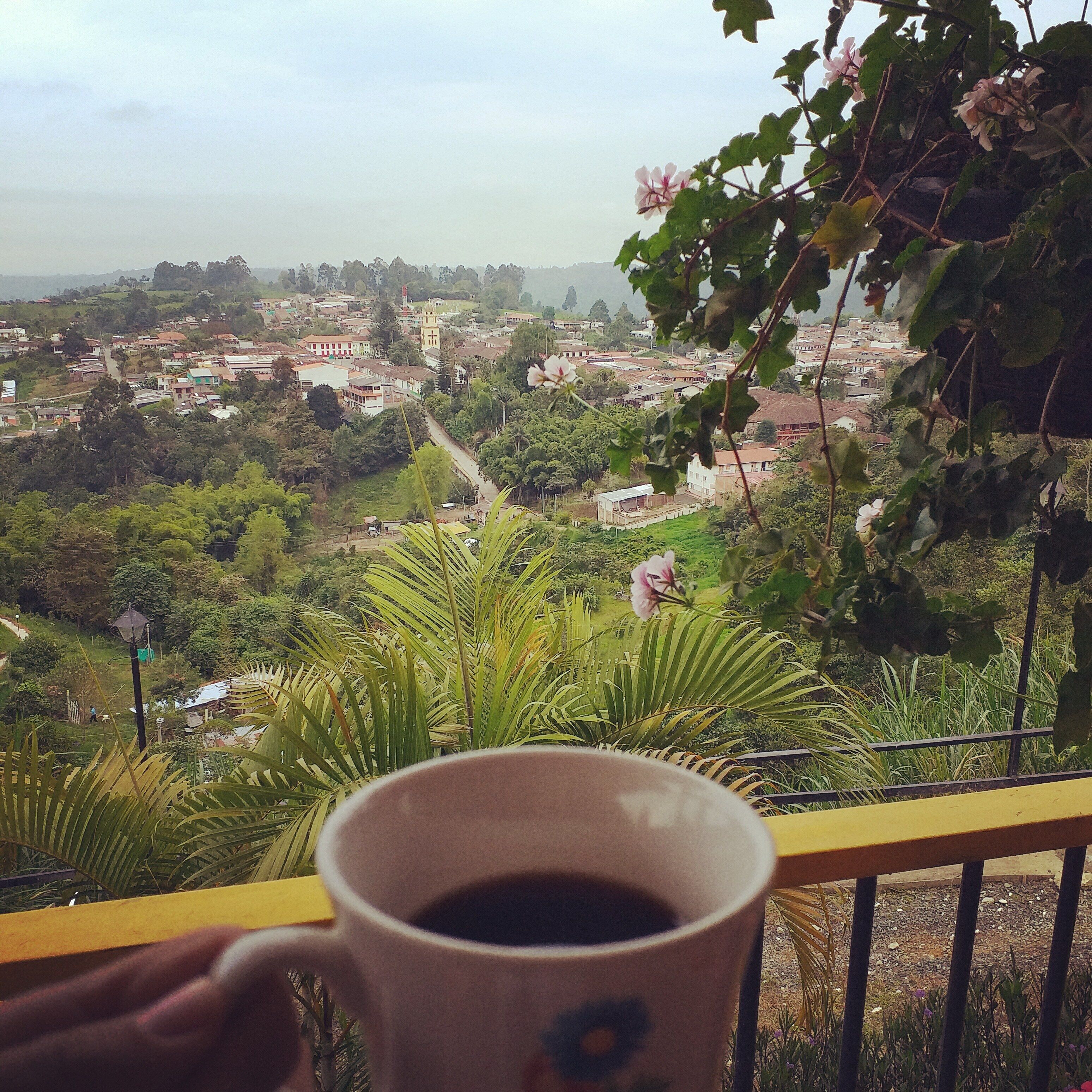 A mug of coffee on a balcony overlooking a town with greenery and buildings.