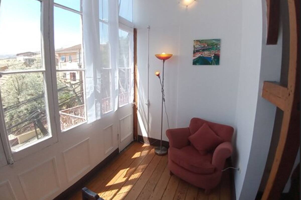 Una habitación con ventanas amplias, una silla roja, una lámpara de pie y un cuadro en la pared.