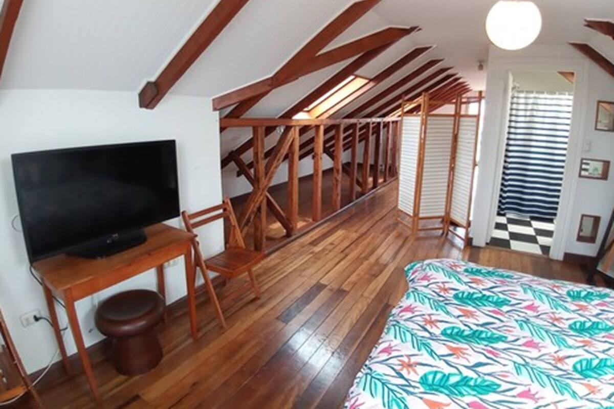 Un dormitorio con techo de madera, una cama con colcha estampada de motivos tropicales, un escritorio de madera con un televisor y una escalera.