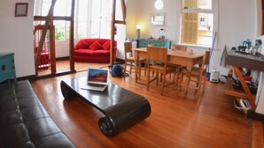 Una habitación con piso de madera, un sofá rojo, una mesa de comedor con sillas, una computadora portátil sobre un tapete y una ventana con persianas.
