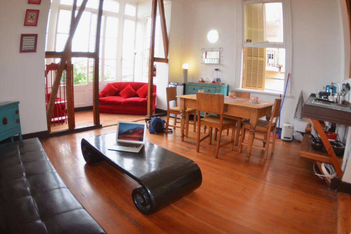 Una habitación con piso de madera, un sofá rojo, una mesa de comedor con sillas, una computadora portátil sobre un tapete y una ventana con persianas.