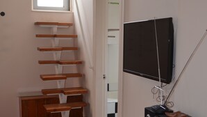 Una habitación con una escalera de madera, un televisor sobre un mueble bajo y una pequeña mesa con altavoces.