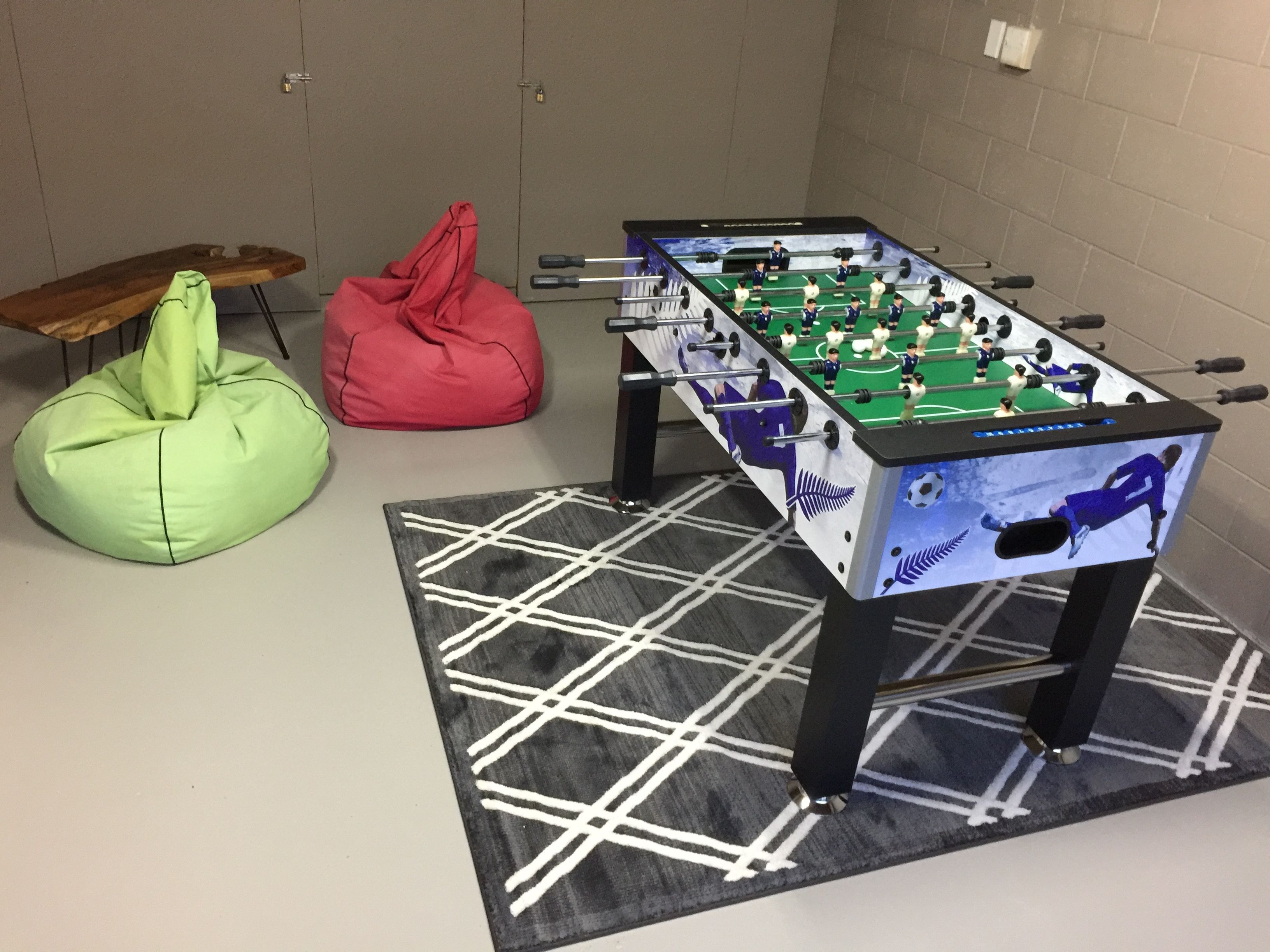 Een tafelvoetbaltafel met een voetbalthema, een groene relaxfauteuil en een rode relaxfauteuil in een kamer met een geruit vloertje.