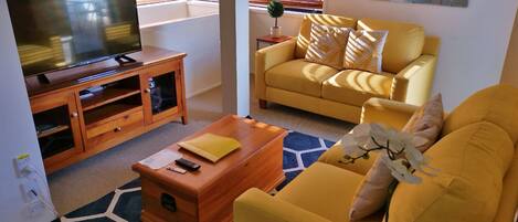 Un salon avec un téléviseur posé sur un meuble en bois, un canapé jaune, une table basse et un tapis bleu à motifs.