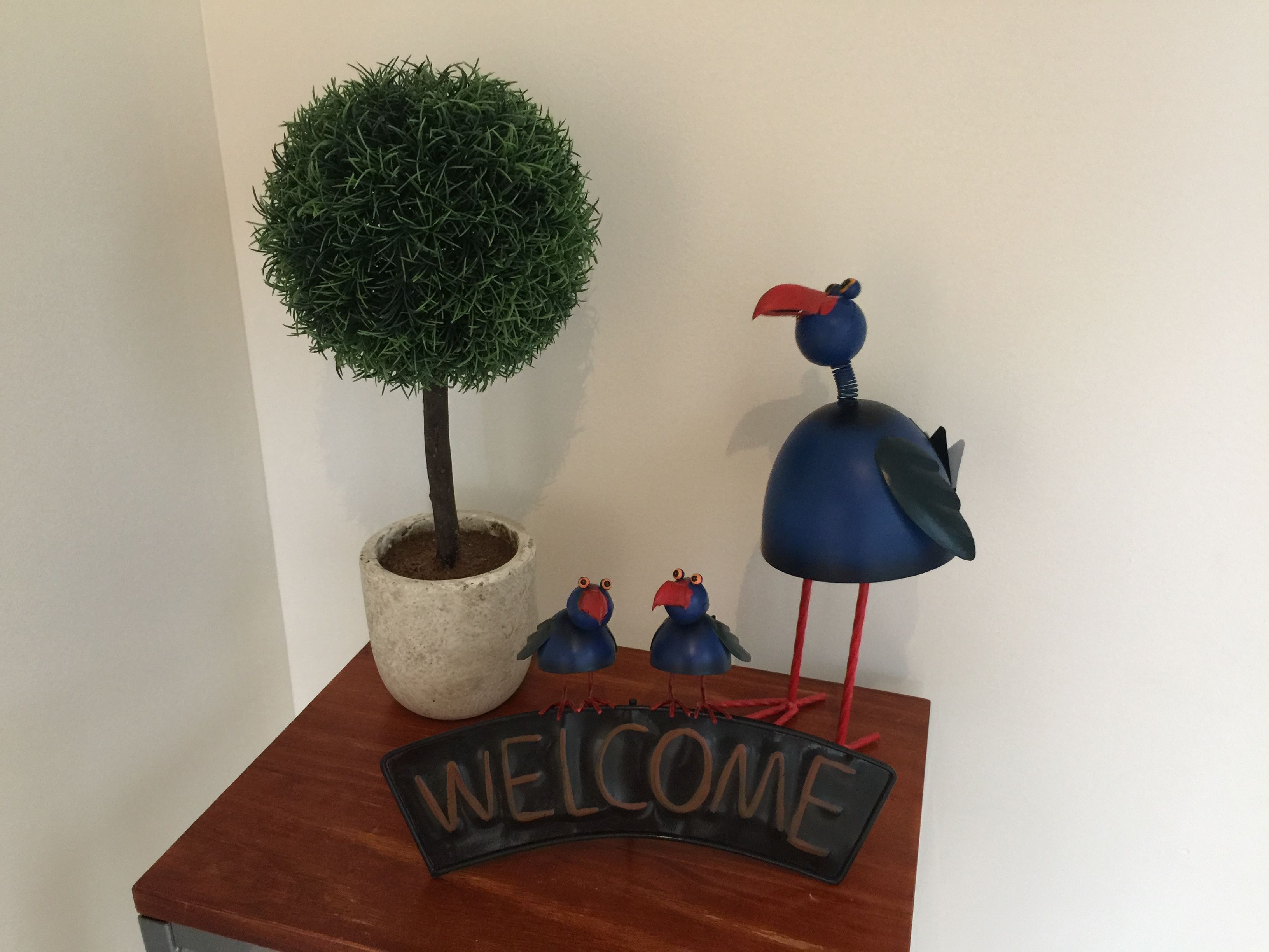 Un letrero de bienvenida, un árbol en maceta y dos esculturas de aves sobre una mesa de madera.