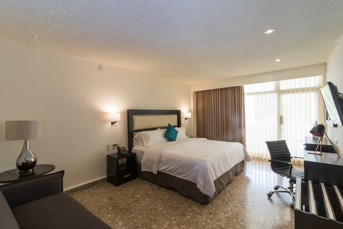 A hotel room with a bed, bedside tables, a desk, a chair, and a large window with curtains.