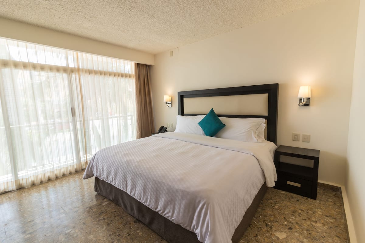 A hotel room with a large bed, a nightstand, a lamp, and a view through sheer curtains.