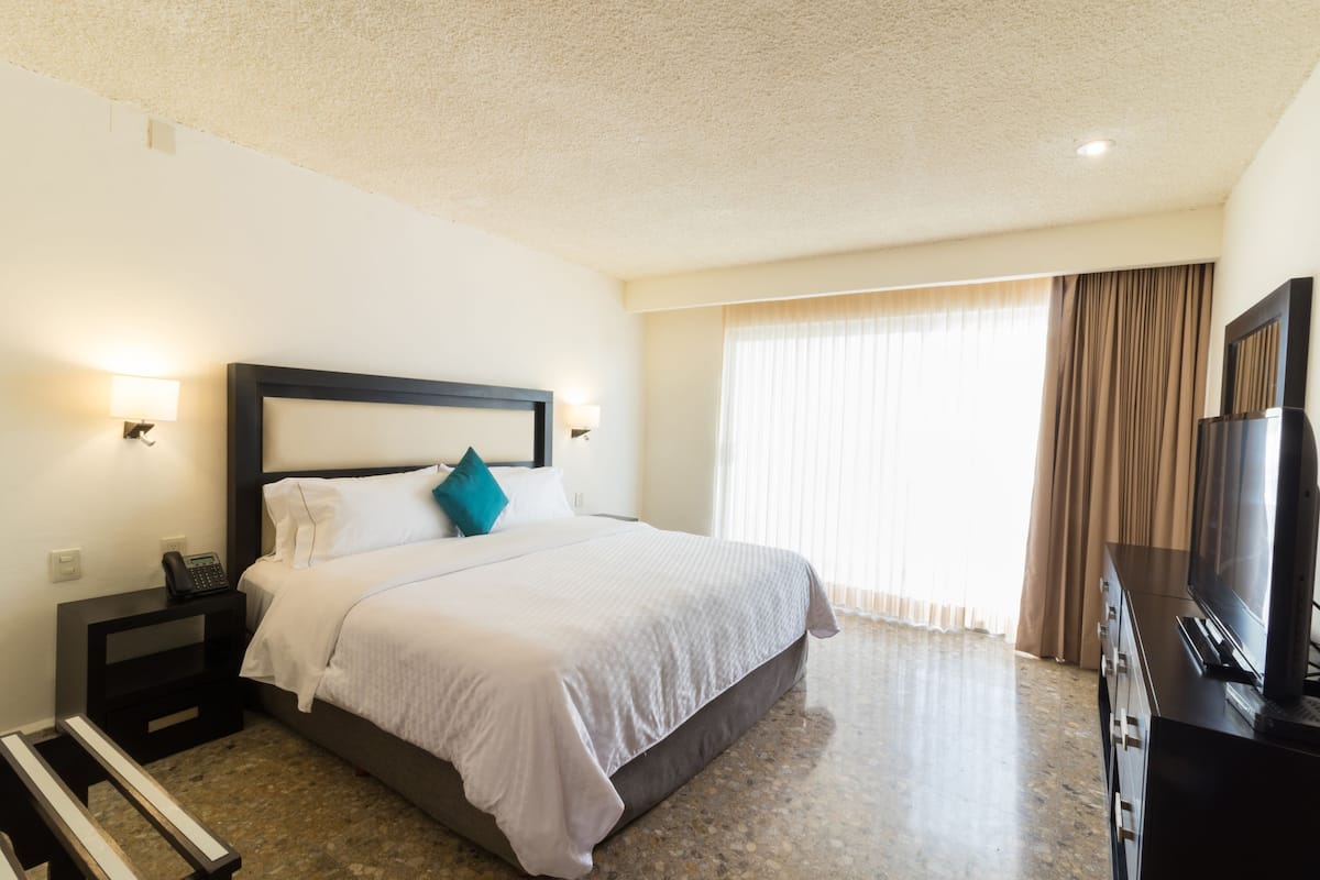 A hotel room with a bed, a TV, a desk, and a window with curtains.