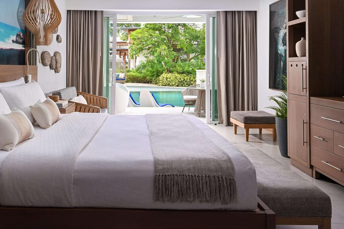 A bedroom with a large bed, a TV, a wardrobe, and a view of a swimming pool and greenery.