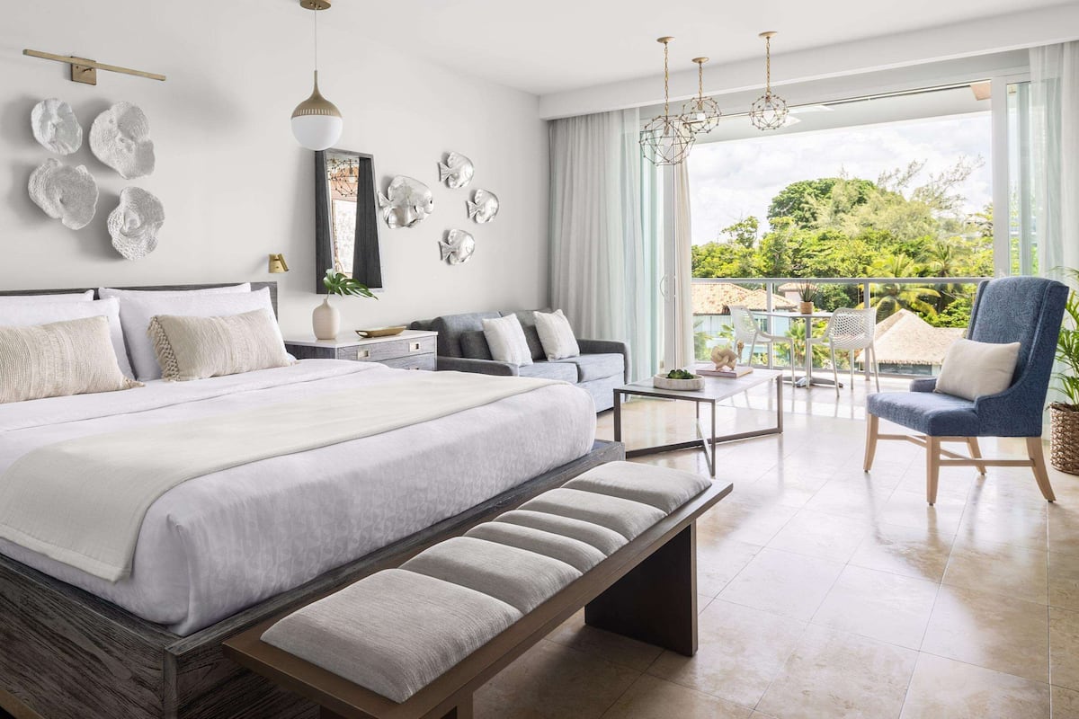 A modern bedroom with a large bed, bedside tables, a bench, a balcony with a chair, and decorative wall art.