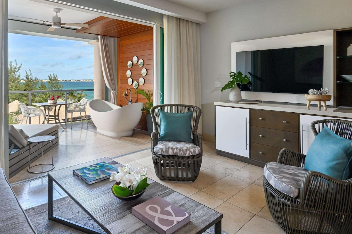 A modern living room with a sofa, coffee table, wicker chairs, a flat-screen TV, and a balcony with a view of the sea.