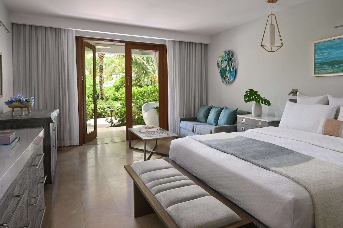 A modern hotel room with a large bed, a desk, a chair, and a balcony with a view of greenery.