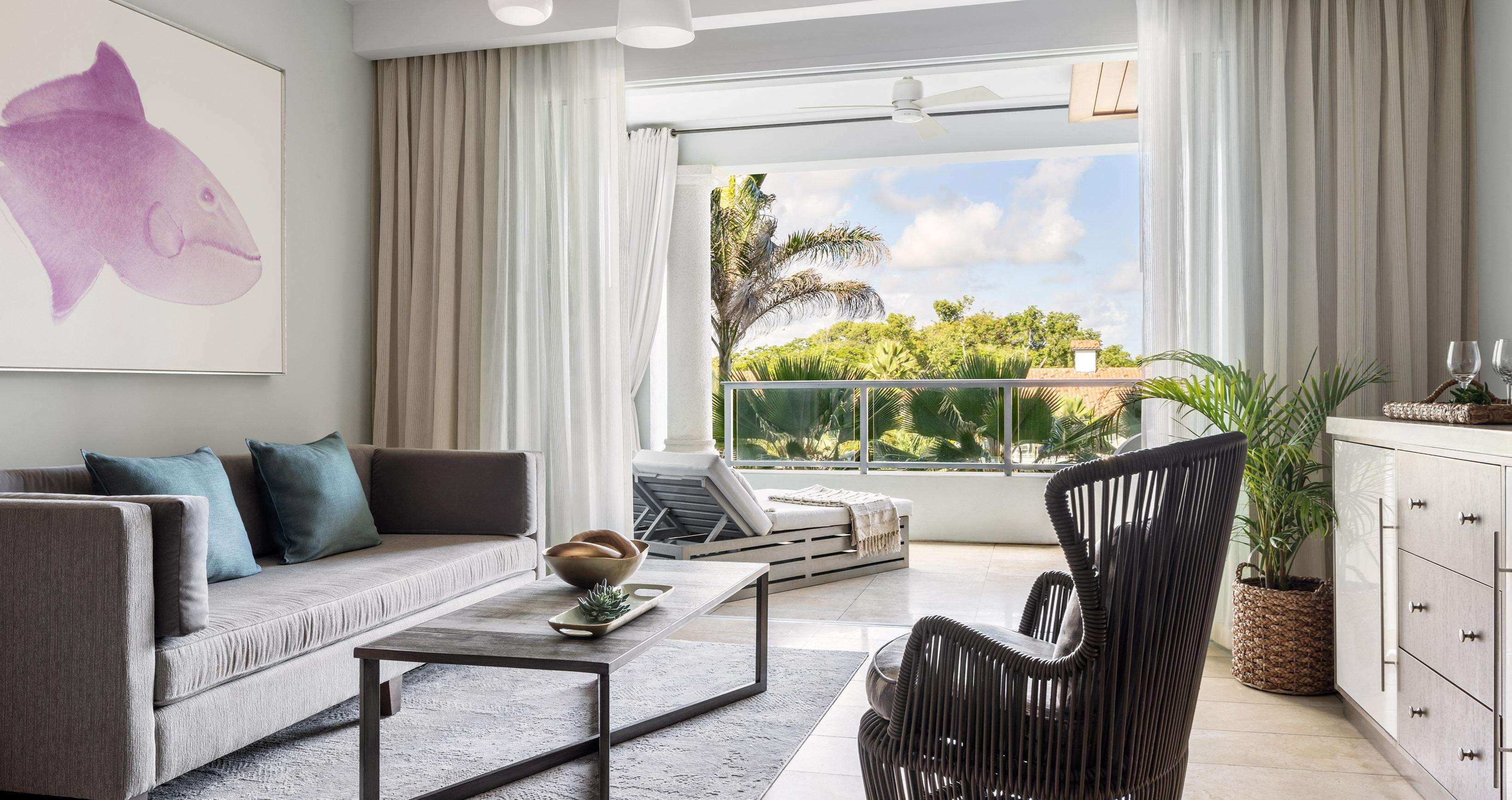 A modern living room with a sofa, coffee table, and a chair. There is a large window with a view of greenery and a building.