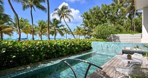 Zona de piscina con agua cristalina, rodeada de exuberante vegetación y palmeras, que da a una playa.