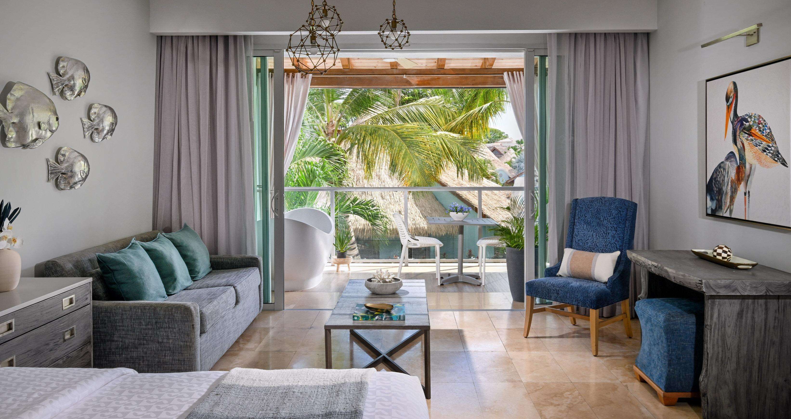 Ein modernes Hotelzimmer mit einer Couch, einem Sessel, einem kleinen Tisch und Blick auf einen tropischen Garten durch große Fenster.