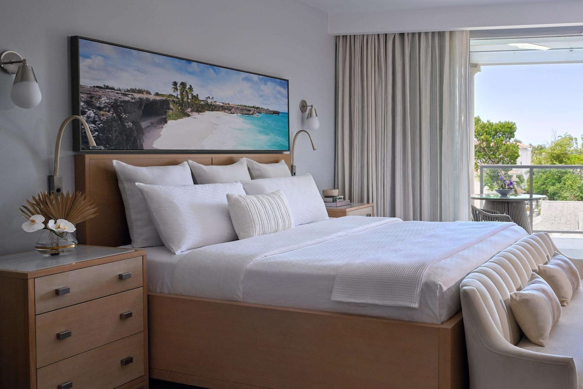 A modern hotel room with a large bed, a view of the sea, a bathtub, and a balcony with a table and chairs.