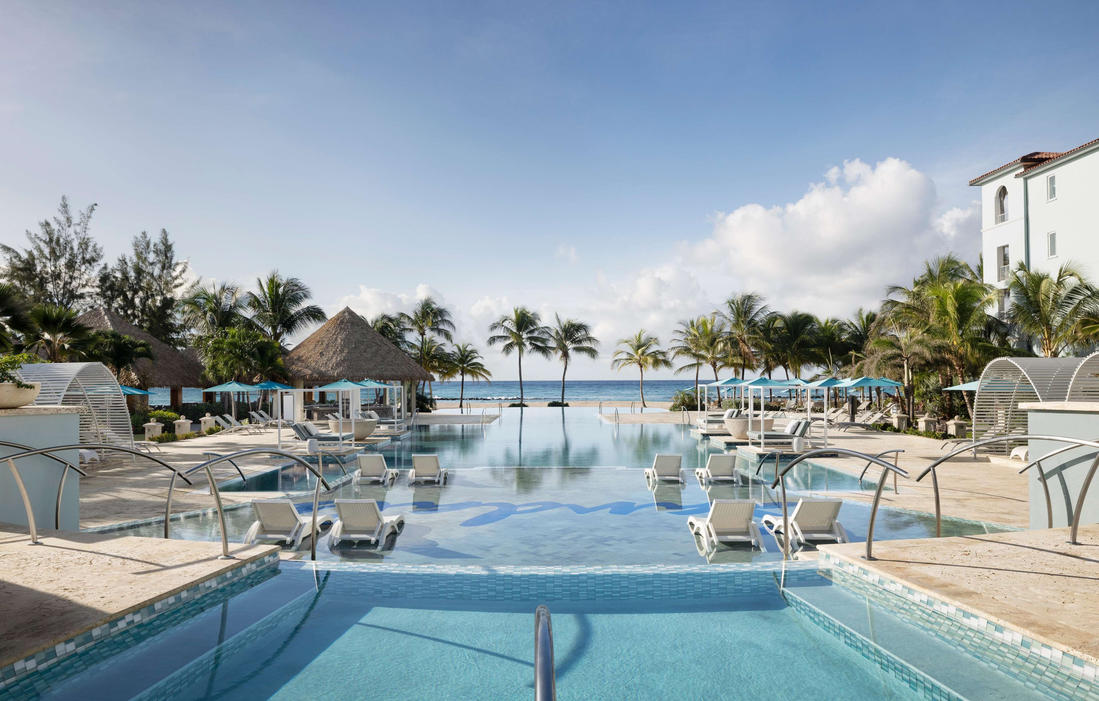 A resort pool with lounge chairs, palm trees, and a view of the ocean.