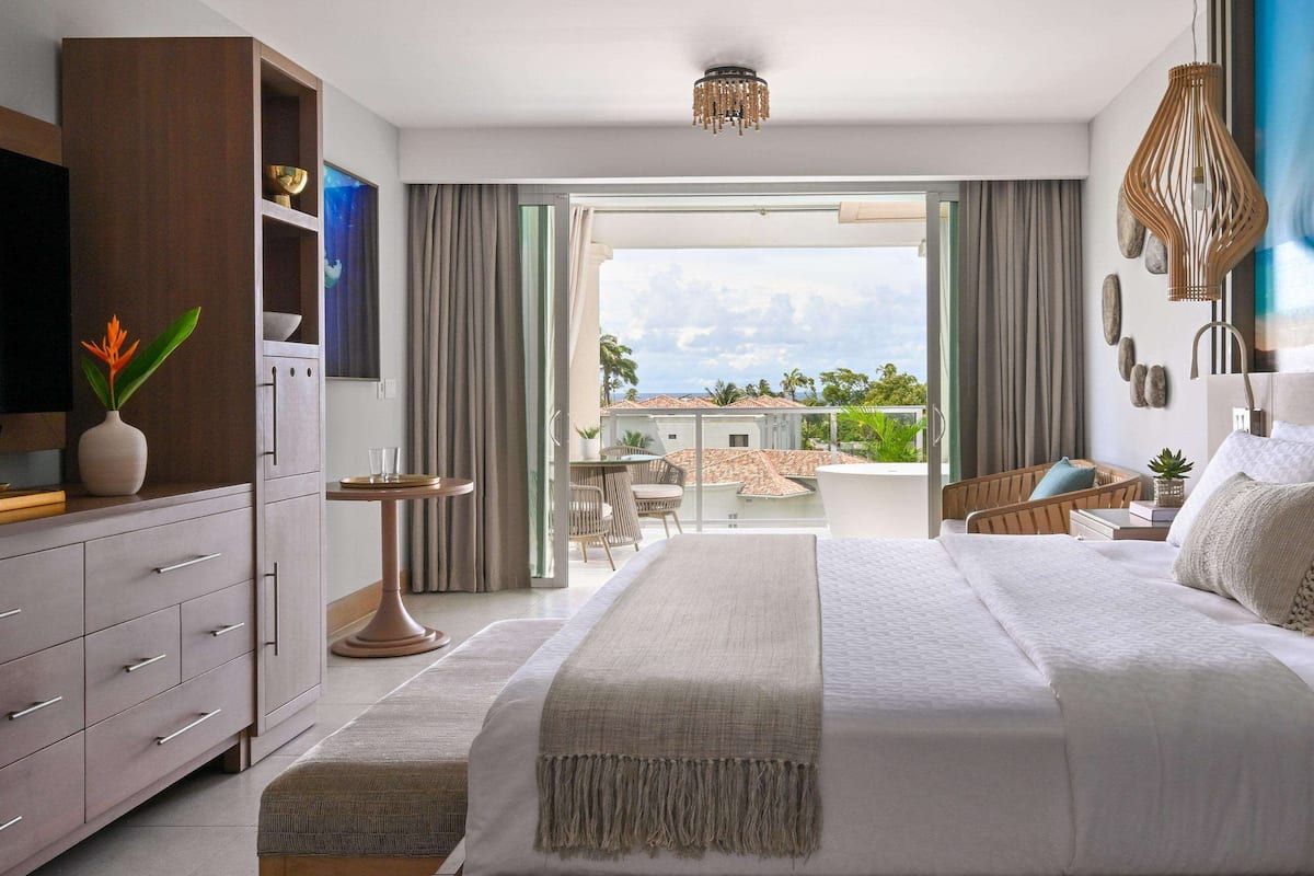 A modern bedroom with a large bed, a TV, a desk, and a balcony with a view of palm trees.