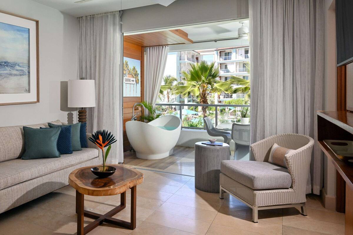 A modern living room with a sofa, armchair, and round wooden table. A painting is on the wall, and there is a view of a balcony with palm trees.