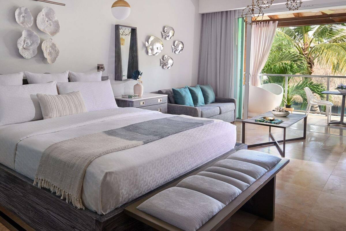 A modern bedroom with a large bed, bedside tables, a bench, and a balcony with a view of palm trees.