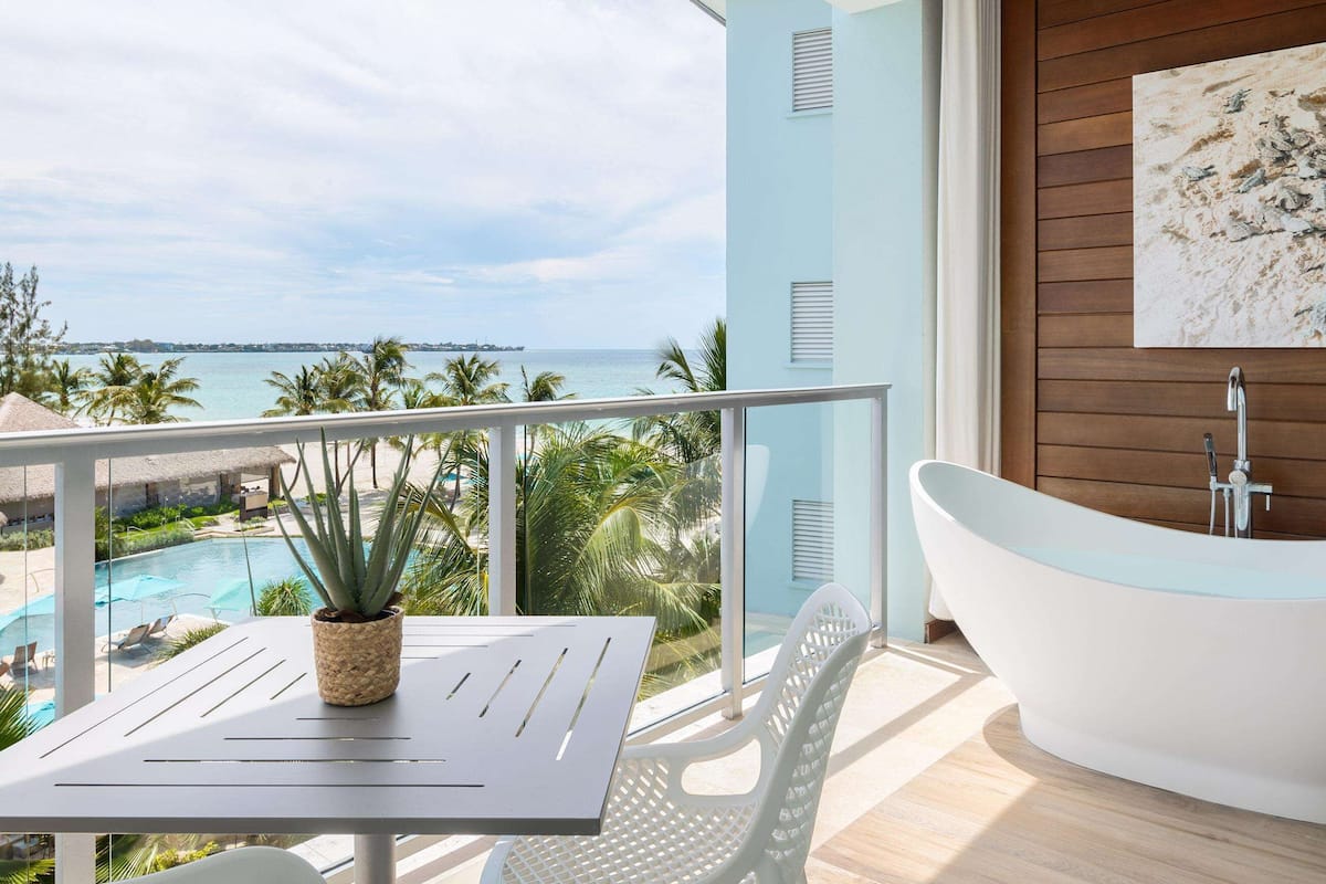A modern bathroom with a freestanding bathtub, a wooden panel wall, and a large window overlooking a pool and palm trees.
