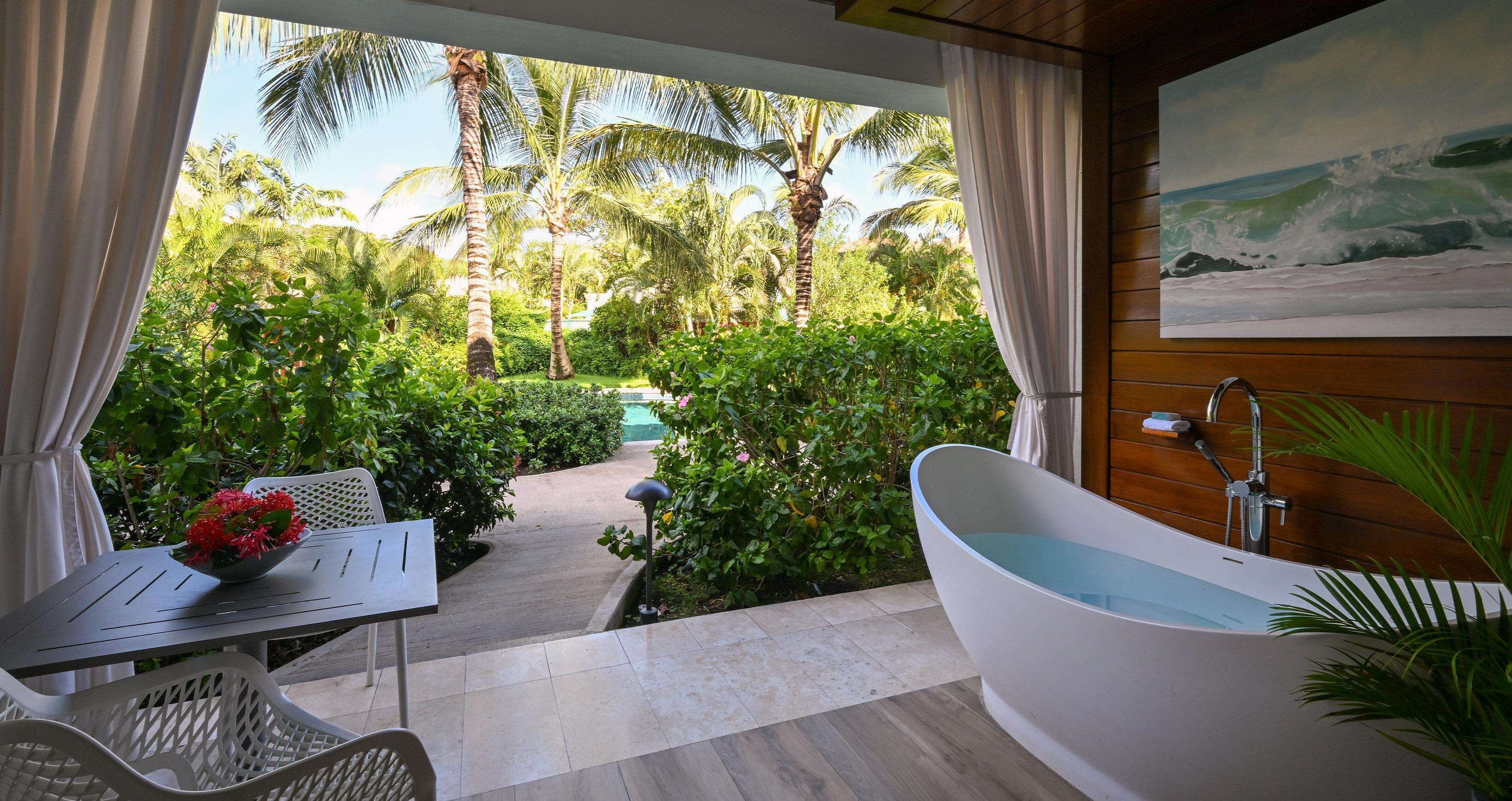 A bathroom with a bathtub, a table with a flower vase, and a view of a tropical garden with palm trees.