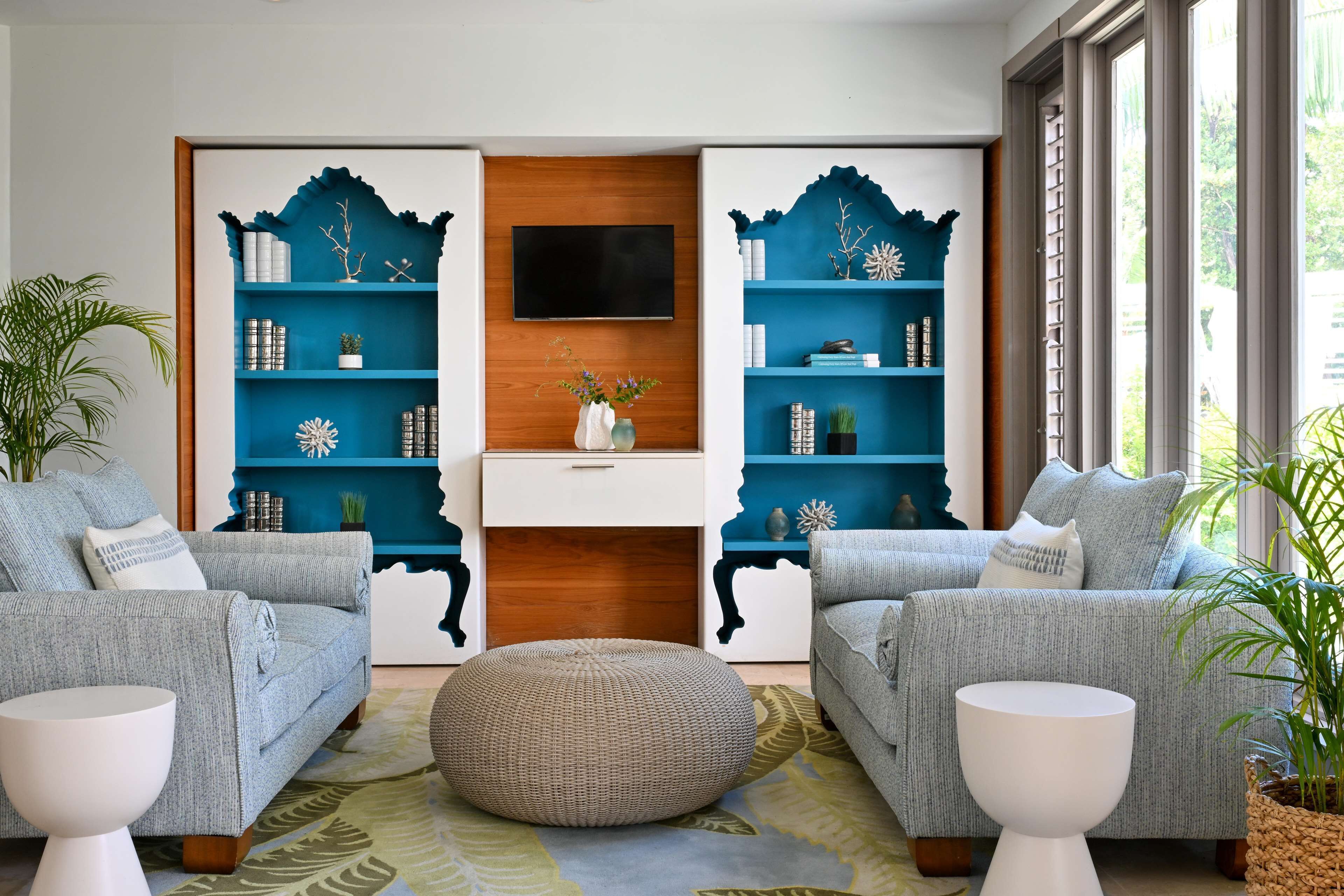 A modern living room with two grey sofas, a white coffee table, and a large woven ottoman. There are two blue bookshelves with decorative items and a flat-screen TV mounted on the wall.