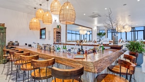 Un área de bar moderna con taburetes de madera, lámparas colgantes de mimbre y un amplio ventanal con vistas al cielo.