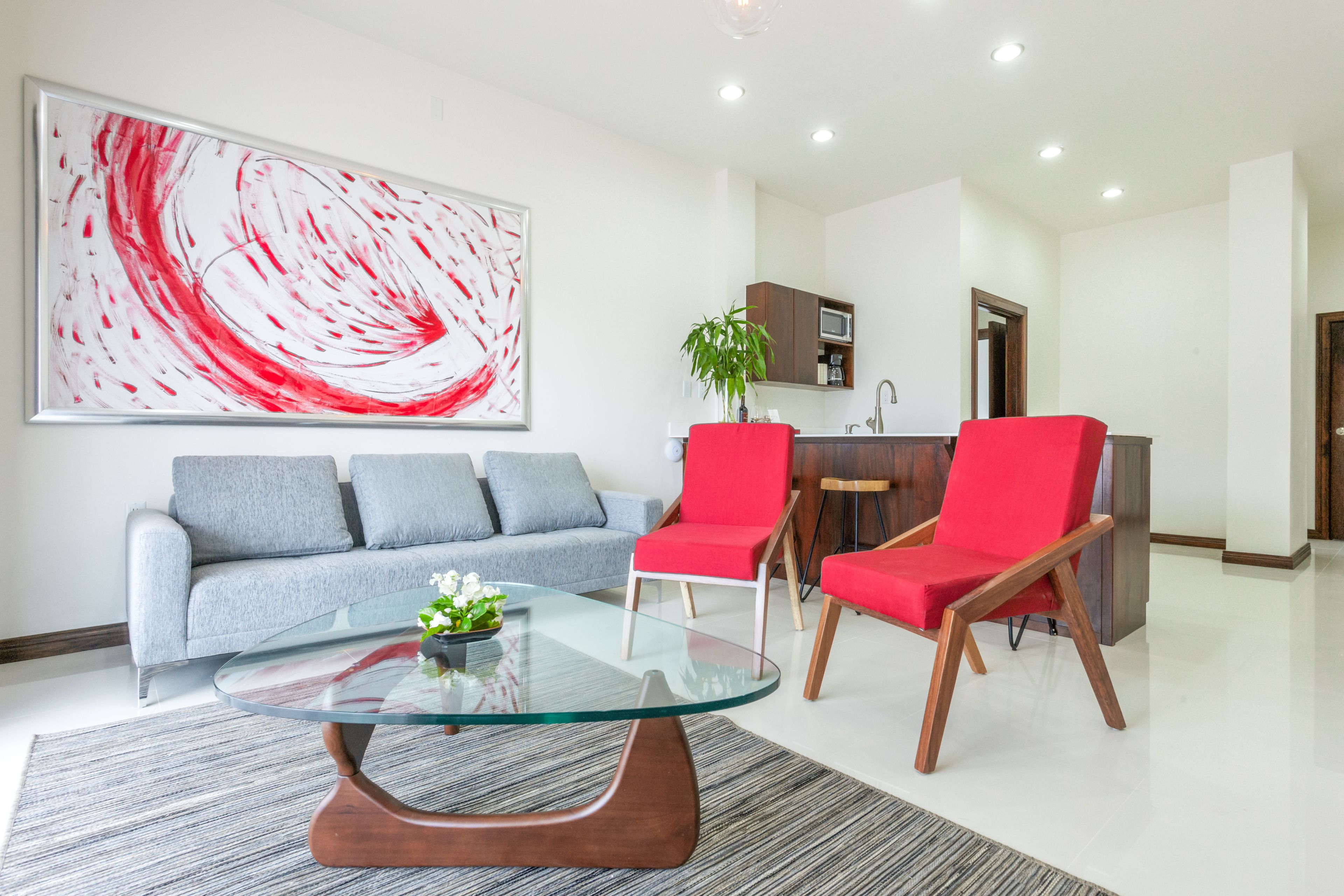 A modern living room with a glass coffee table, grey sofa, red chairs, and a large abstract painting.