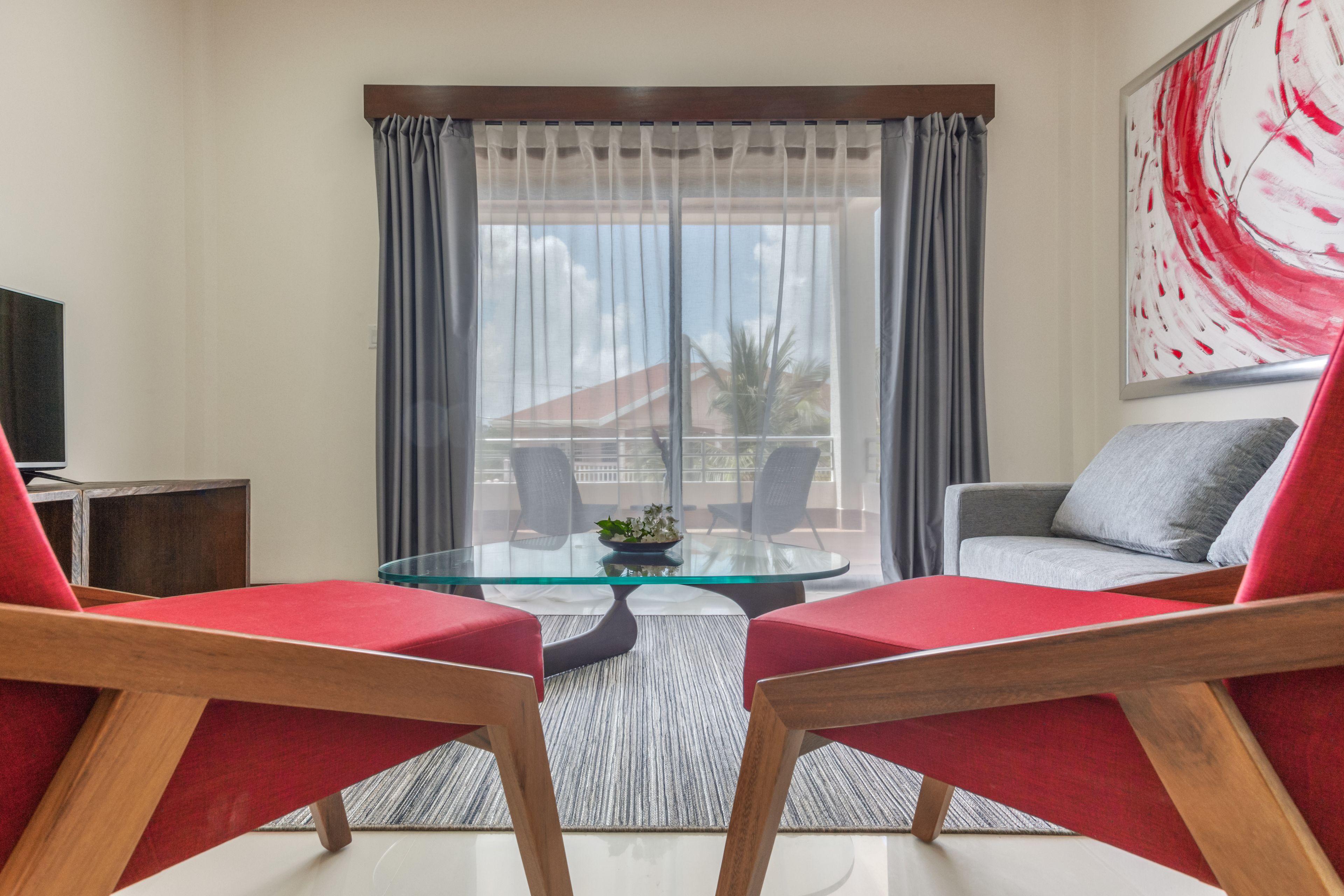 A modern living room with a glass table, red and grey chairs, a sofa, and a large window with curtains.